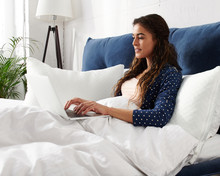 Happy casual beautiful woman working on a laptop sitting on the bed in the house