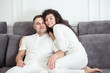 © uv_group - Portrait of young couple sitting together on grey sofa in modern white apartment or domestic room. Happy lovely family embracing and hugging together close-up