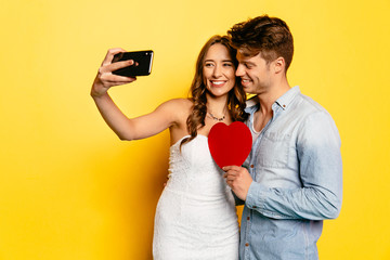 Cheerful woman taking selfie on smartphone with her attractive boyfriend holding red heart. St. Valentine's day