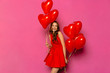 Smiling girl holding air balloons in two hands, posing at camera. St. Valentine's day. Dressed in red dress, on pink background.