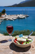 A glass of red wine and bowl of greek salad with greek flag on by the sea view, summer greek holidays concept.