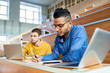 © Seventyfour - Portrait of two students sitting at desks in modern auditorium at college and preparing for class, focus on young Middle-Eastern man writing in copybook
