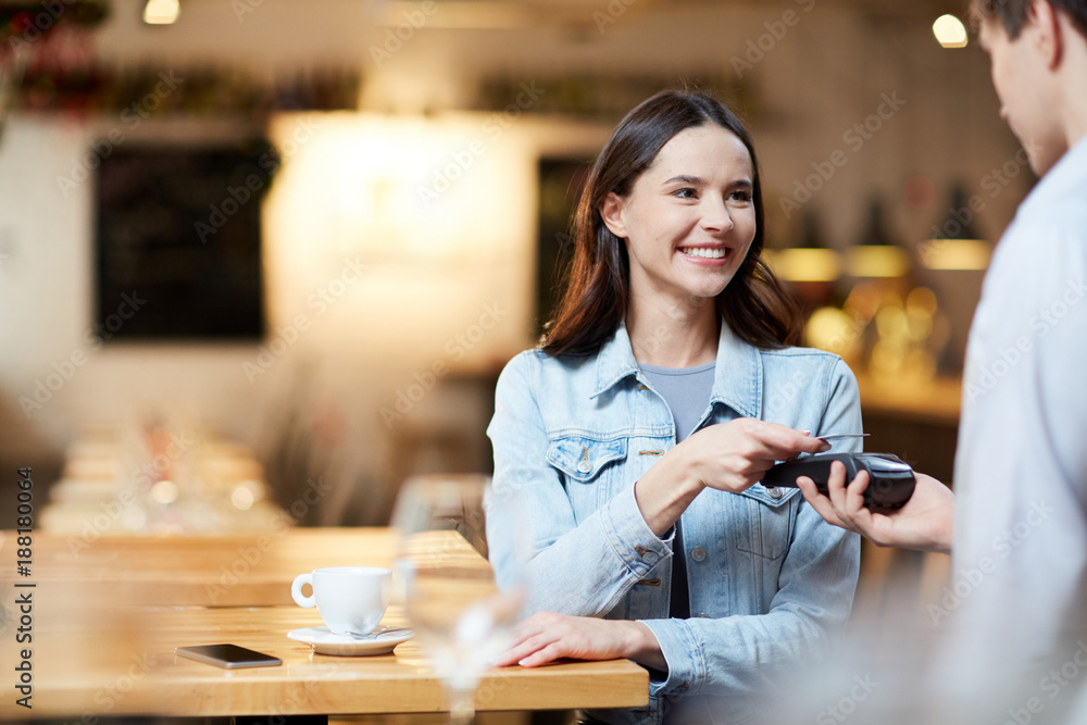 Young smiling woman with card holding it over payment terminal while paying for her order in cafe - obrazy, fototapety, plakaty 