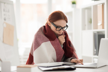 Frozen young manager wrapped in plaid sitting by workplace in front of laptop and working in the net