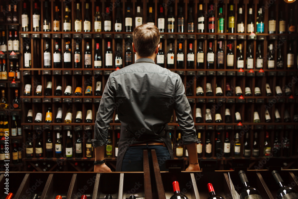 Bartender at wine cellar full of bottles with exquisite drinks - obrazy, fototapety, plakaty 