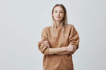 Wall Mural - Waist-up portrait of beautiful student girl with blonde straight hair smiling cheerfully while listening to compliments, wearing loose long-sleeved sweater, keeping arms folded. Beauty and youth