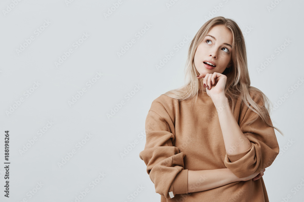 Portrait of pensive fair-haired dreamy hipster girl imagines something in her mind, looks upwards, being deep in thoughts, touching chin. Attractive young blonde female dreams about weekends - obrazy, fototapety, plakaty 
