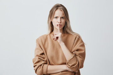 Wall Mural - Blonde attractive european female looks at camera keeps finger on lips, being displeased and asks not to make noise, poses in studio. Horizontal shot of woman frowning face, making hush gesture