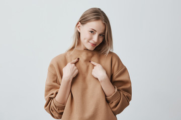 Wall Mural - Shot of beautiful attractive blonde woman smiling, pointing with index fingers at herself, dressed in beige long-sleeved sweater, expressing positive emotions and feelings. Beauty and youth concept