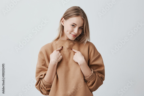 Fotomural Shot of beautiful attractive blonde woman smiling, pointing with index fingers at herself, dressed in beige long-sleeved sweater, expressing positive emotions and feelings