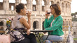Two happy young woman tourists sitting at the table of a bar restaurant in front of the Colosseum in Rome drink and toast with a glass of italian red wine. Stylish colorful dress on a summer day at