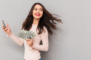 Poster - Portrait of happy brunette female 30s winning lots of money dollar currency using her smartphone rejoicing about salary or prize over gray wall