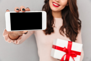 Poster - Cropped portrait of successful brunette female 30s rejoicing her new silver smartphone holding present box over gray wall