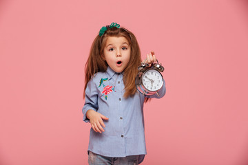 Wall Mural - Shot of female kid posing on camera with eyes and mouth wide open holding clock nearly 6 being shocked or shaken up over pink background