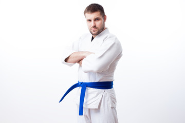 Young sporty man with dark hair in a white kimono with blue belt for sambo, jiu jitsu, judo stands with his arms crossed on a white isolated background