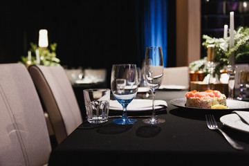 Close up glasses, forks, knives, napkins, candles and decorative flower on dark black tablecloth on table served for dinner in cozy restaurant. Empty served restaurant table with plates and wine glass