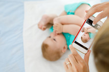 Parent Taking Photo Of A Baby With Smartphone. Adorable Newborn Child Taking Foot In Mouth