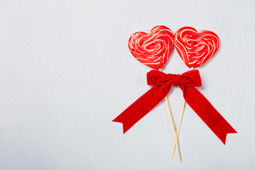 Coffee for Valentine`s day morning in white cup with two red heart lollipops and red ribbon bow on grey or blue background, copy space top view, creamy froth