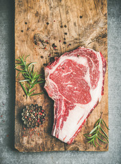 Wall Mural - Flat-lay of raw prime beef meat dry-aged steak rib-eye on bone over rustic wooden board over grey concrete countertop background with seasoning, top view, copy space. Meat high-protein dinner concept