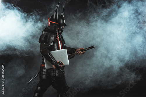 Photo  side view of samurai in traditional armor with laptop taking out sword on dark b