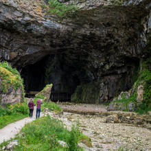 Smoo Cave, Durness, Scotland. A Tourist Couple Adding Scale To The Scene As They Photograph The Entrance To Smoo Cave In Northern Scotland.