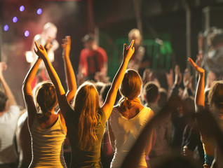 Audience with hands raised at a music festival and lights streaming down from above the stage.