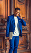 © Alexander Image - African American Businessman with little goatee working in New York, wearing blue suit, white collarless shirt, holding laptop computer, standing by vintage office doorway, talking on cell phone..