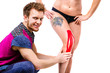 © Elizaveta - the young doctor the therapist sets on the knee a beautiful strong with a good figure of the patient kinesiology tape of red color. The concept of sports medicine and recovery from injuries.