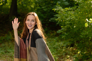 Wall Mural - Young brunette woman in a wool poncho standing in the sun on a footpath in a Park green and waving in greeting.