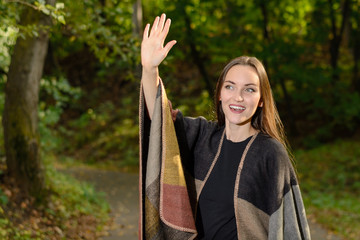 Wall Mural - Young brunette woman in a wool poncho standing in the sun on a footpath in a Park green and waving in greeting.