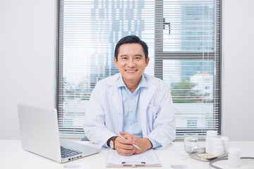 Wall Mural - Smiling asian doctor sitting at his desk in medical office