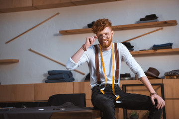 young male fashion designer in eyeglasses with measuring tape around neck smiling at camera at workplace