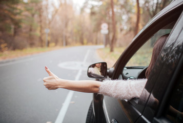 Female traveler on the road holding thumbs up. Fall vacation, holidays, travel, road trip and people concept. Close up Hand. Autumn concept. Autumn forest journey by car