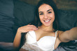 © estradaanton - sensual young woman in white underwear on black lingerie making selfie using a smartphone and smiling while lying on bed.