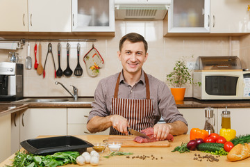 Wall Mural - Attractive caucasian young man in apron sitting at table with vegetables, cooking at home preparing meat stake from pork, beef or lamb, in light kitchen with wooden surface, full of fancy kitchenware.