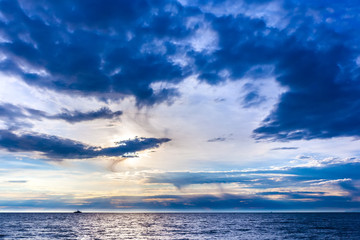 The blue beautiful landscape of the sky with graceful clouds blockaded the bright sun over the sea, the ship in the distance on the horizon. Beautiful colorful sunset on the Black Sea, Sochi, Russia.