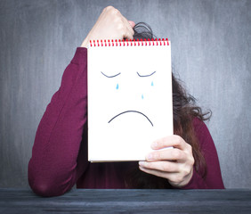 Wall Mural - Woman holding a notebook with drawn sad face, depression concept.