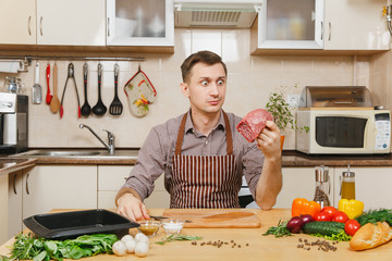 Wall Mural - Attractive caucasian young man in apron sitting at table with vegetables, cooking at home preparing meat stake from pork, beef or lamb, in light kitchen with wooden surface, full of fancy kitchenware.