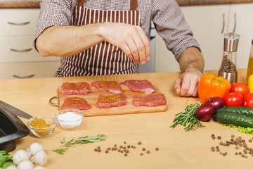 Wall Mural - Close up young man in apron sitting at table with vegetables, cooking at home preparing meat stake from pork, beef or lamb, salt meat, in light kitchen with wooden surface, full of fancy kitchenware.