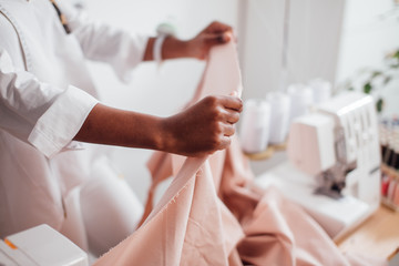 Young african woman seamstress looking at pink fabrics and standing in workshop