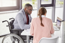 Serious Dark Skinned Male Invalid In Wheelchair Collaborates With Young Female, Make Business Plan Together, Watch Online Conference Or Webinar Online, Sit In Spacious Cabinet. Disabled Person