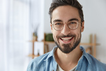 Close Up Shot Of Cheerful Satisfied Attractive Male With Stubble, Has Broad Smile, Wears Round Spectacles, Rejoices Success At Work, Stands Against Cozy Interior. Fashionable Designer Glad Be Praised