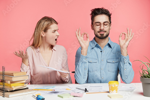 Fotografie, Obraz Angry irritated pretty female student looks at male partner who meditates at working table, tries to relax for continuing hard work on course paper, isolated over pink background