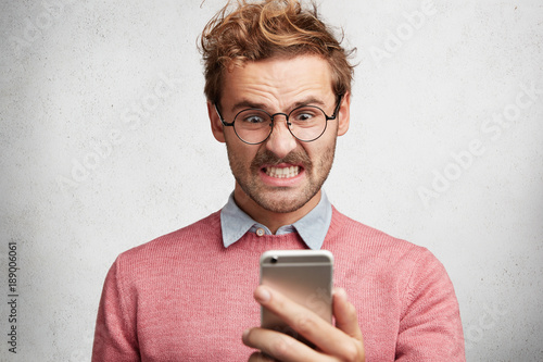 Fotografía Angry irritated male reads text message on smart phone remind him about bills, n