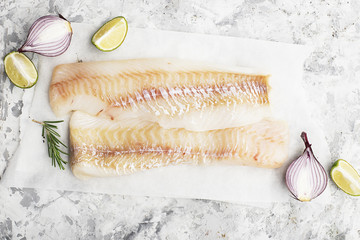 Fillet of fresh raw sea cod on a sheet of parchment on a simple light background with sea salt, ground pepper, sweet onion rosemary and lime slices before cooking. Top View.