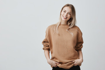 Wall Mural - Portrait of young woman in casual clothes with blonde dyed hair, gently smiling during pleasant conversation, standing in closed posture, keeping arms crossed while talking with friends.