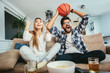 © Mediteraneo - Couple is watching basketball game on the sofa at home.