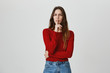 © Cookie Studio - Hush. Portrait of young beautiful caucasian girl with dark long hair in red sweater and denim jeans holding finger in front of lips, looking in camera with serious face expression. Body language