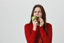 Studio Shot Of Emotional Young Female With Long Hair Dressed In Red Sweater Biting Green Apple And Frowning Face. Cute Student Girl Dressed Casually Has Healthy Snack