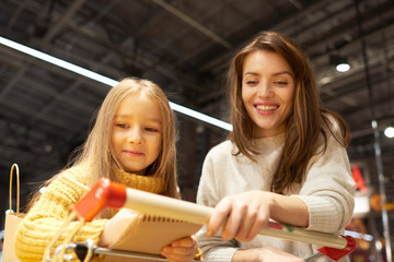 Wall Mural - Portrait of cheerful young woman grocery shopping with daughter in supermarket reading shopping list and leaning on cart